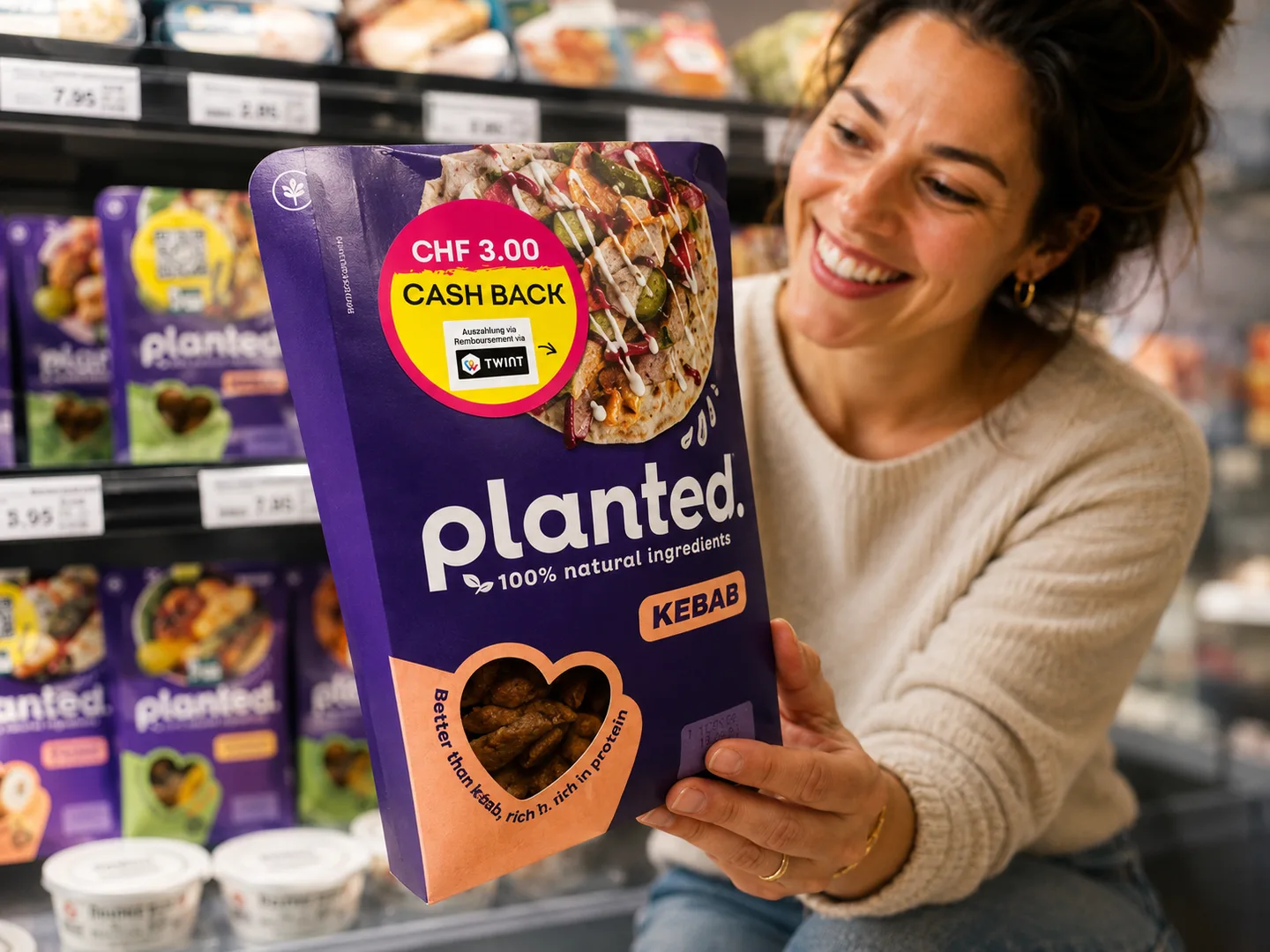 A shopper holds a planted Kebab pack with a CHF 3.00 TWINT cashback sticker at the shelf.